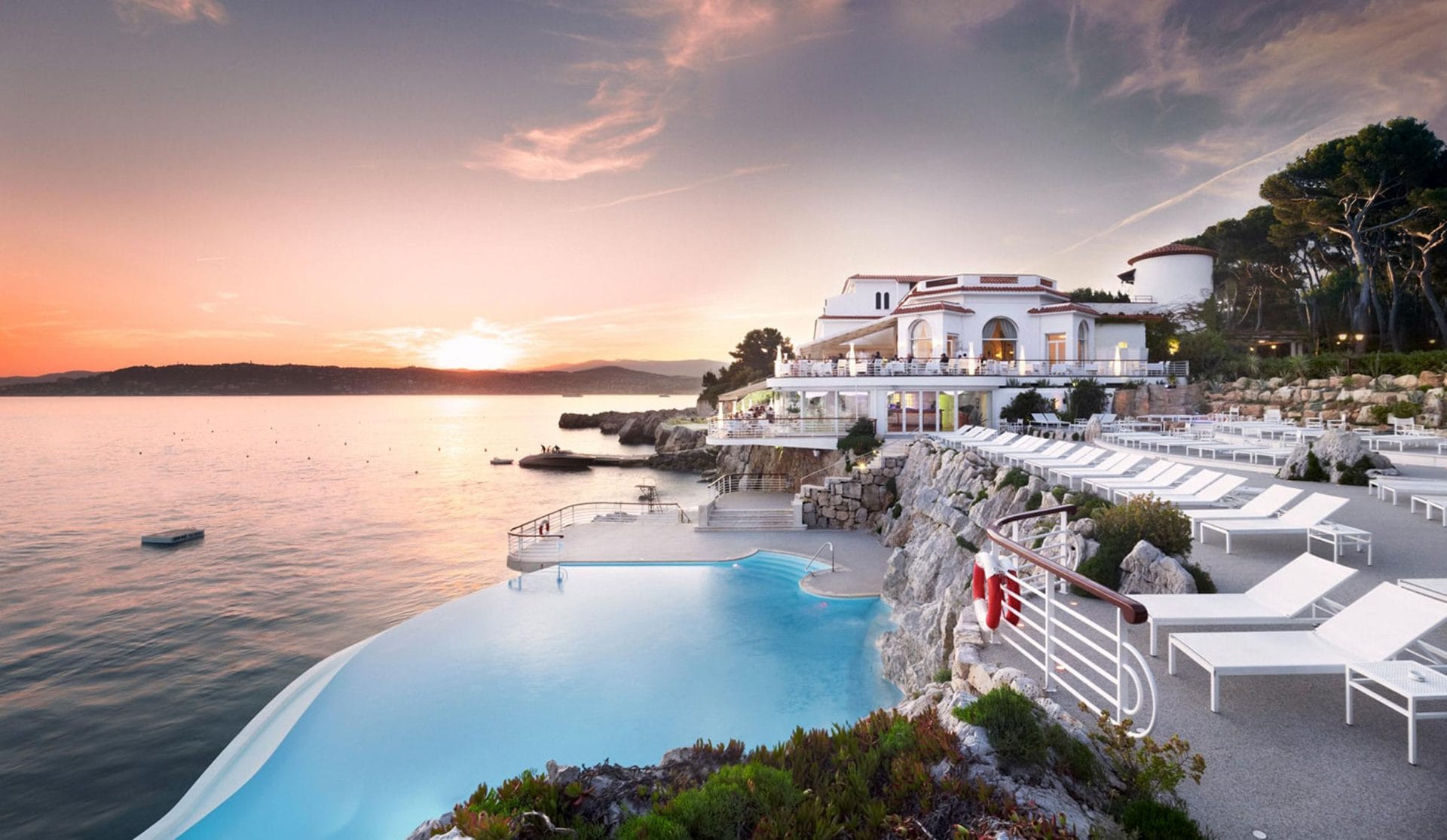 Luxurious seaside resort with infinity pool, white loungers, and setting sun reflecting on a calm ocean horizon.