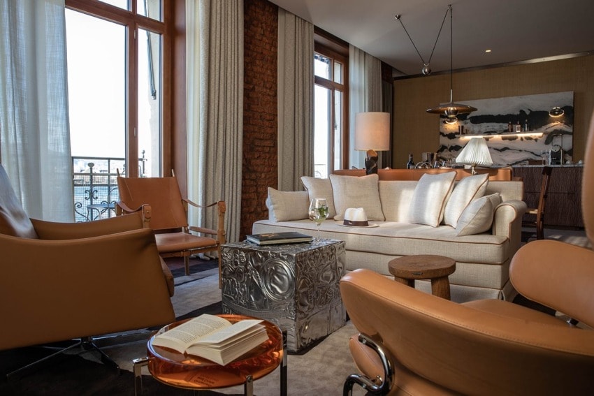 Luxurious hotel suite with modern decor, leather chairs, a sofa, open book, and a city view through large windows.