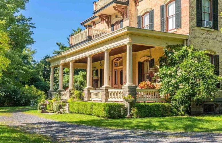 Historic brick house with a large porch, surrounded by greenery and a gravel driveway in a sunny landscape.