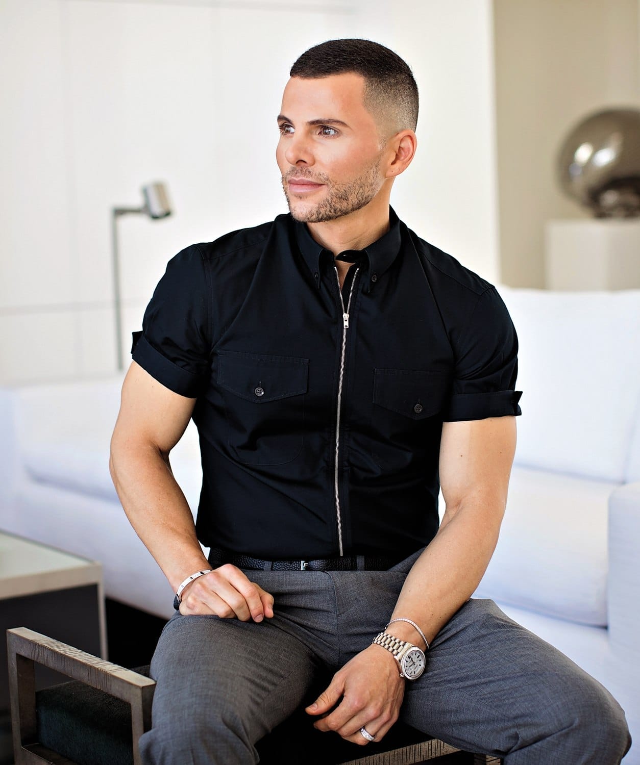 Man in black shirt and gray pants sitting on a chair in a modern room.