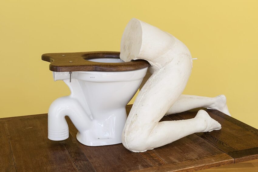 Surreal sculpture of a headless human figure kneeling into a toilet, set against a yellow background.
