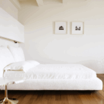 Minimalist bedroom with white bed, wooden floor, and abstract wire chair. Two framed photos on wall.