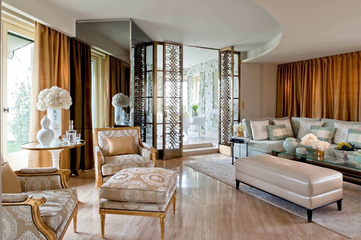 Luxurious living room with elegant furniture, gold accents, mirrored wall, and natural light through large windows.