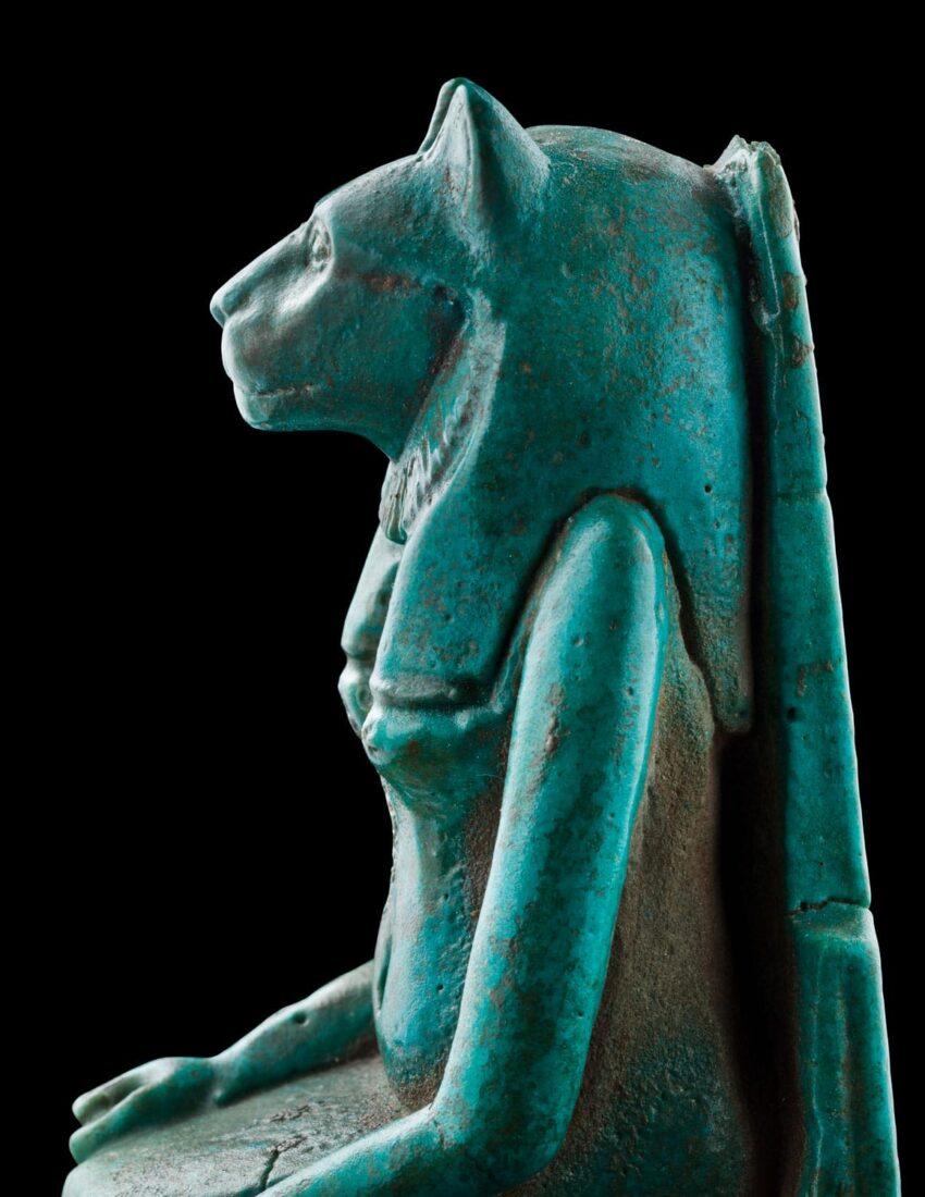 Turquoise statue of an Egyptian deity with a lioness head and human body on a black background.