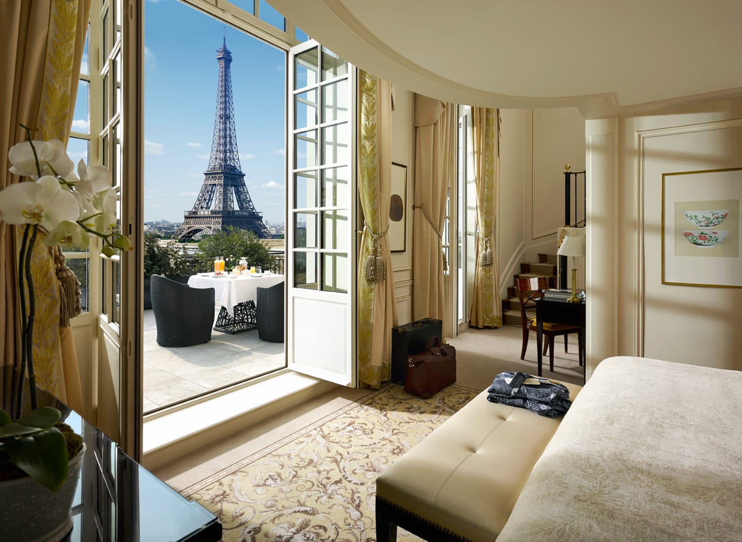 Luxurious hotel room with a large balcony overlooking the Eiffel Tower, set for a meal with open doors and bright decor.