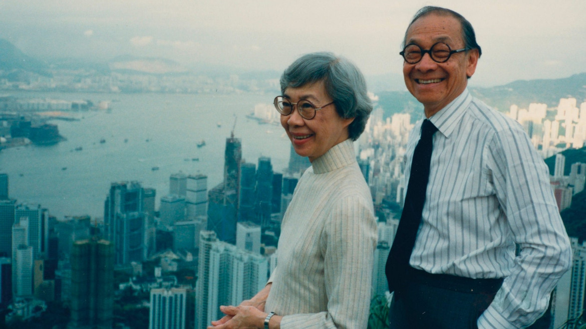 Two people smiling with a city skyline and harbor in the background on a cloudy day.