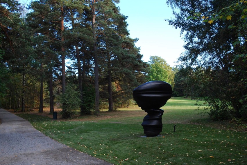Abstract black sculpture in a park surrounded by trees and a grassy field.
