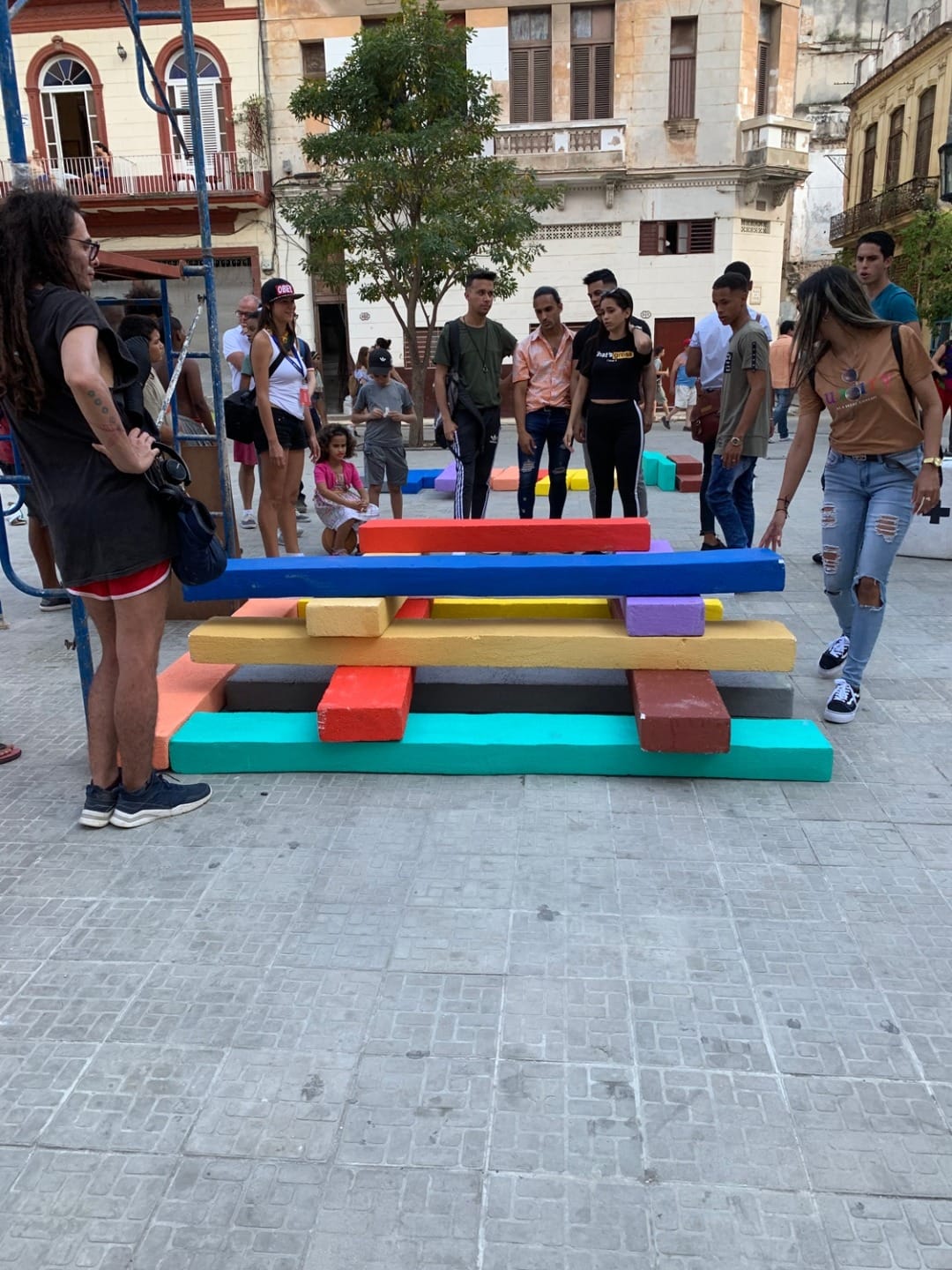 People gathered around a colorful outdoor art installation with stacked rectangular blocks in an urban setting.