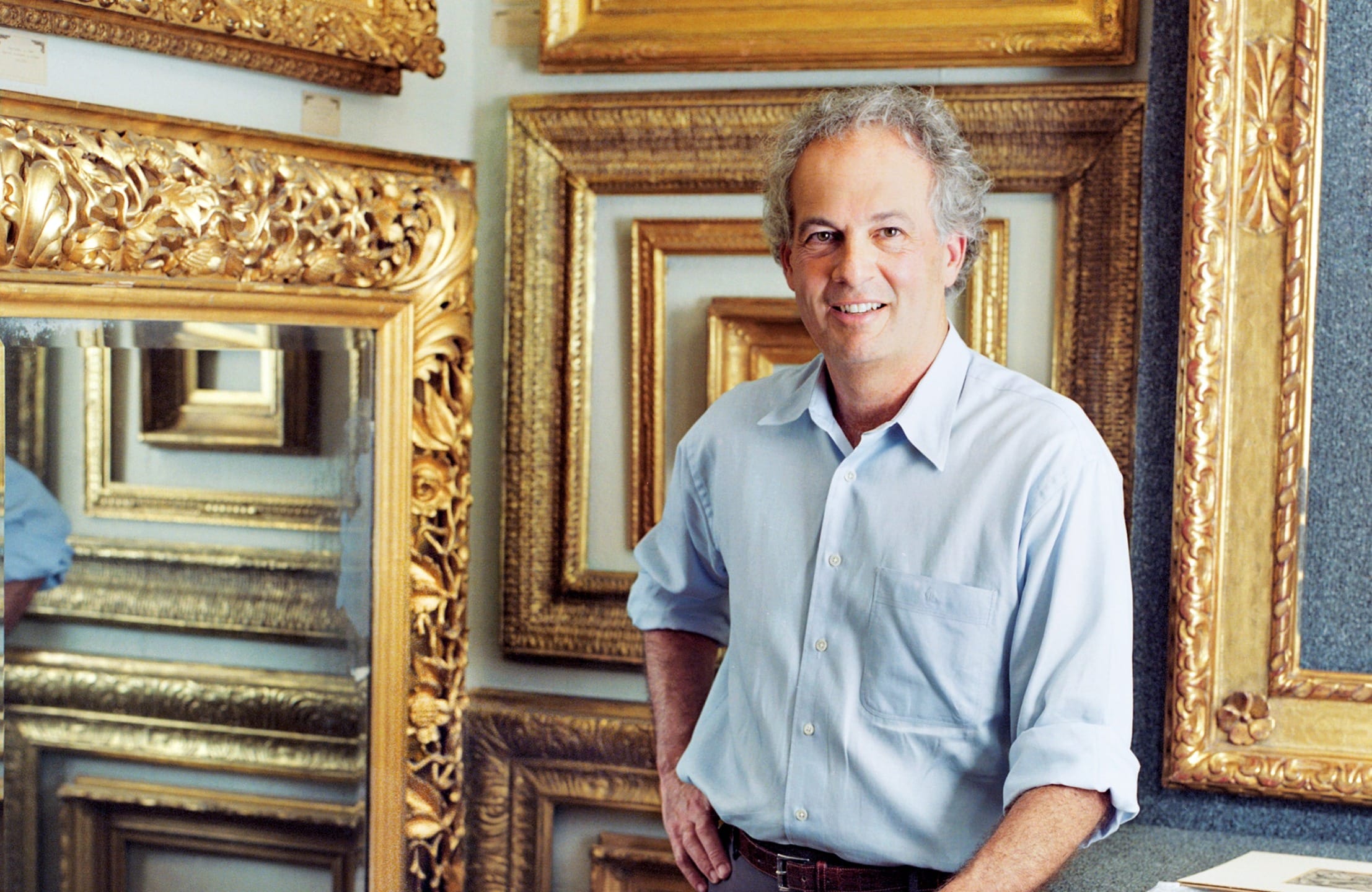 Man in blue shirt standing beside ornate gold frames displayed on a wall.