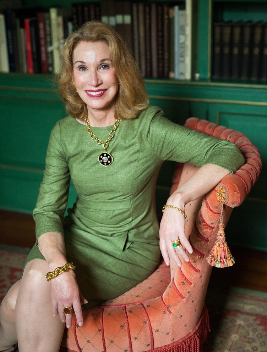 Woman sitting on a striped chair in a green dress with a necklace and bracelets, bookshelves in the background.