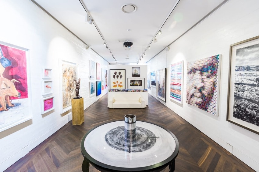 Modern art gallery with diverse paintings and sculptures, featuring a central round table in a well-lit room.