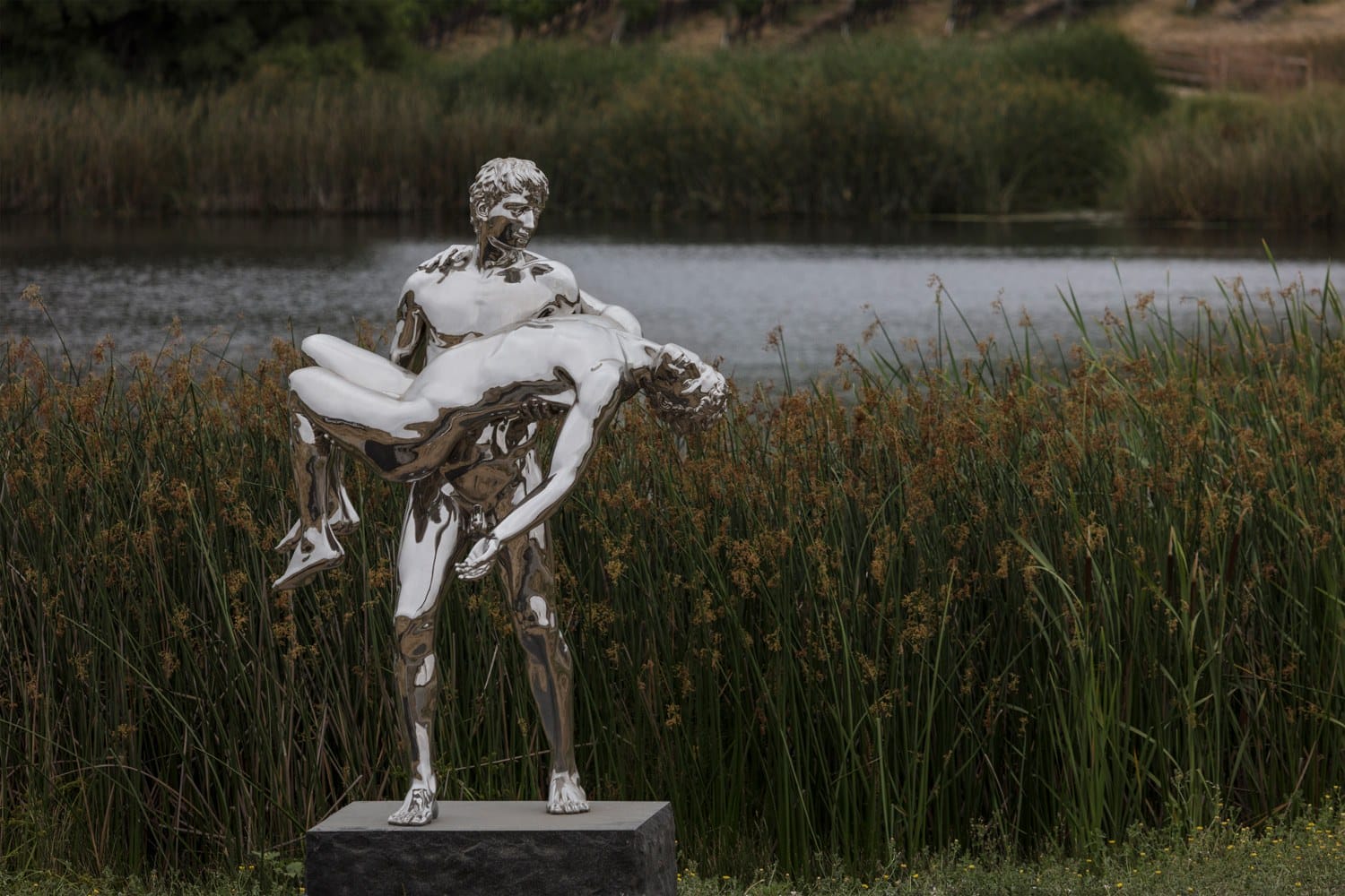 Modern chrome sculpture of two figures by a lake, one carrying the other, set in a natural landscape with tall grasses.
