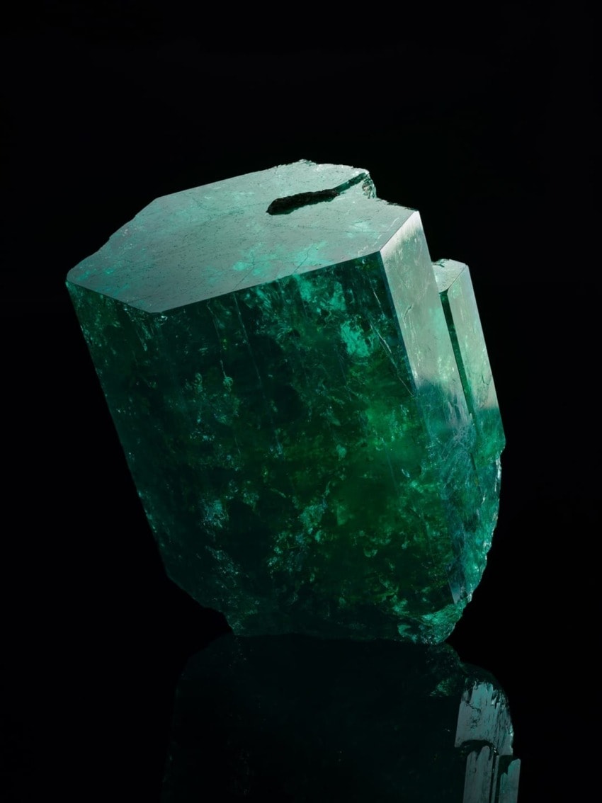 Emerald crystal with hexagonal shape and vibrant green color on a dark reflective surface.