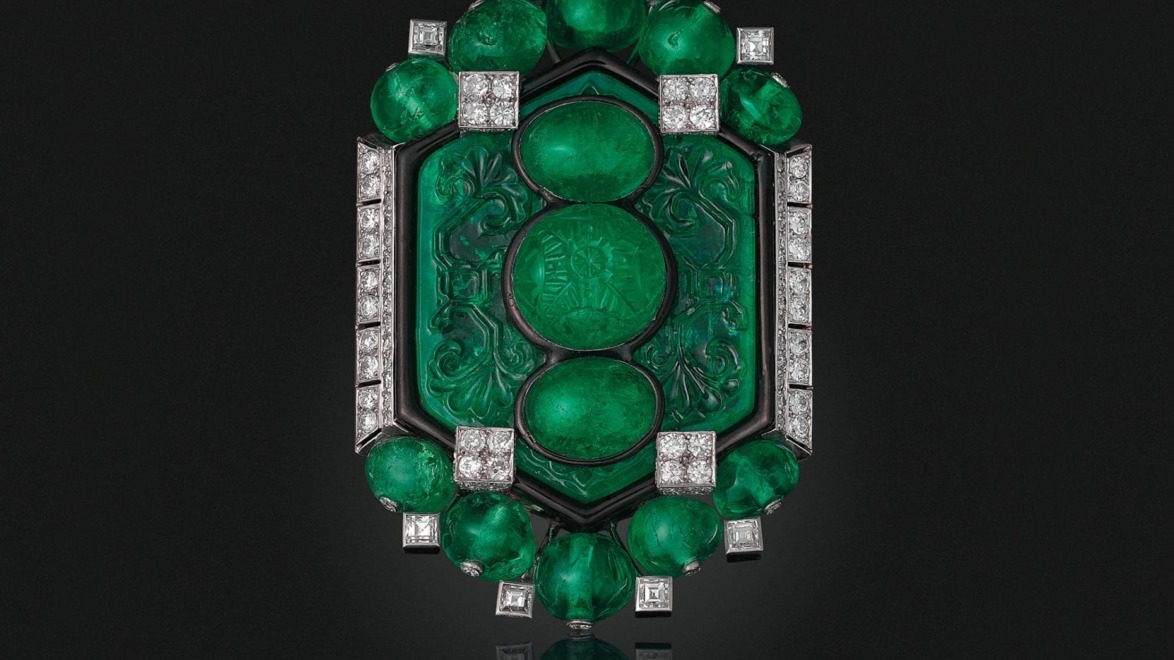 Emerald and diamond Cartier brooch with intricate carvings on a dark background.