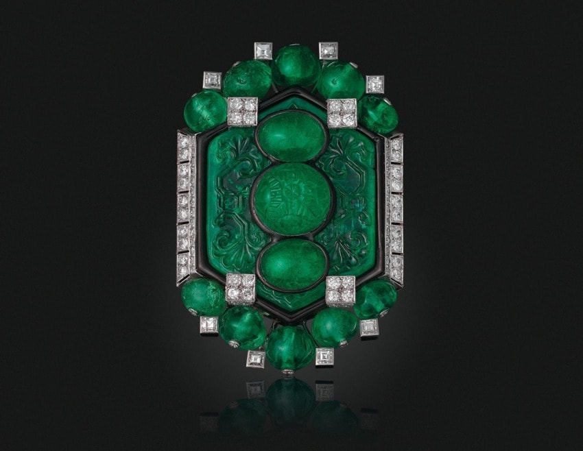 Emerald and diamond Cartier brooch with intricate carvings on a dark background.
