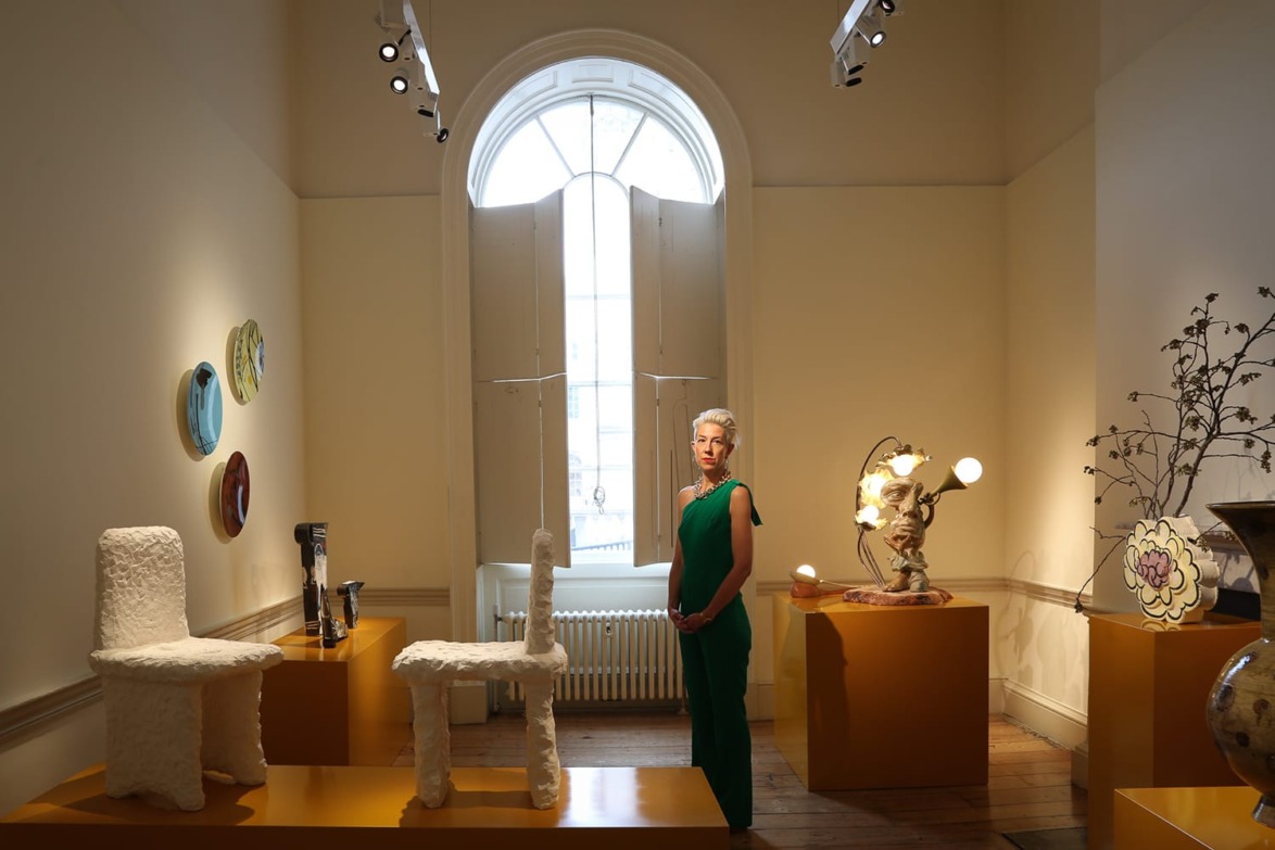 Art gallery with a person in green standing among sculptures and art pieces, large arched window in the background.