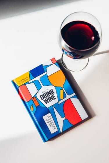 Book titled "How to Drink Wine" with a colorful cover next to a glass of red wine on a white surface.