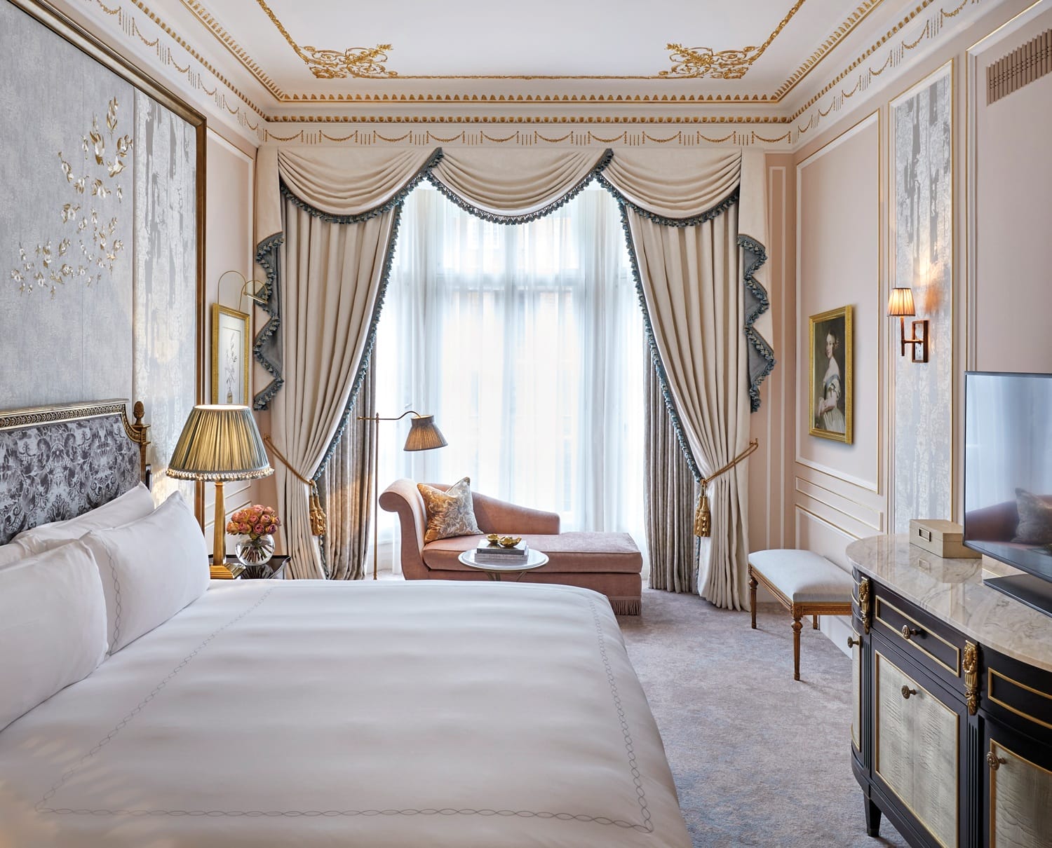 Elegant hotel room with a large bed, classic decor, chaise lounge, and a curtained window letting in natural light.