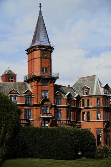 Historic red brick building with a tall spire, numerous windows, and lush greenery in the foreground.