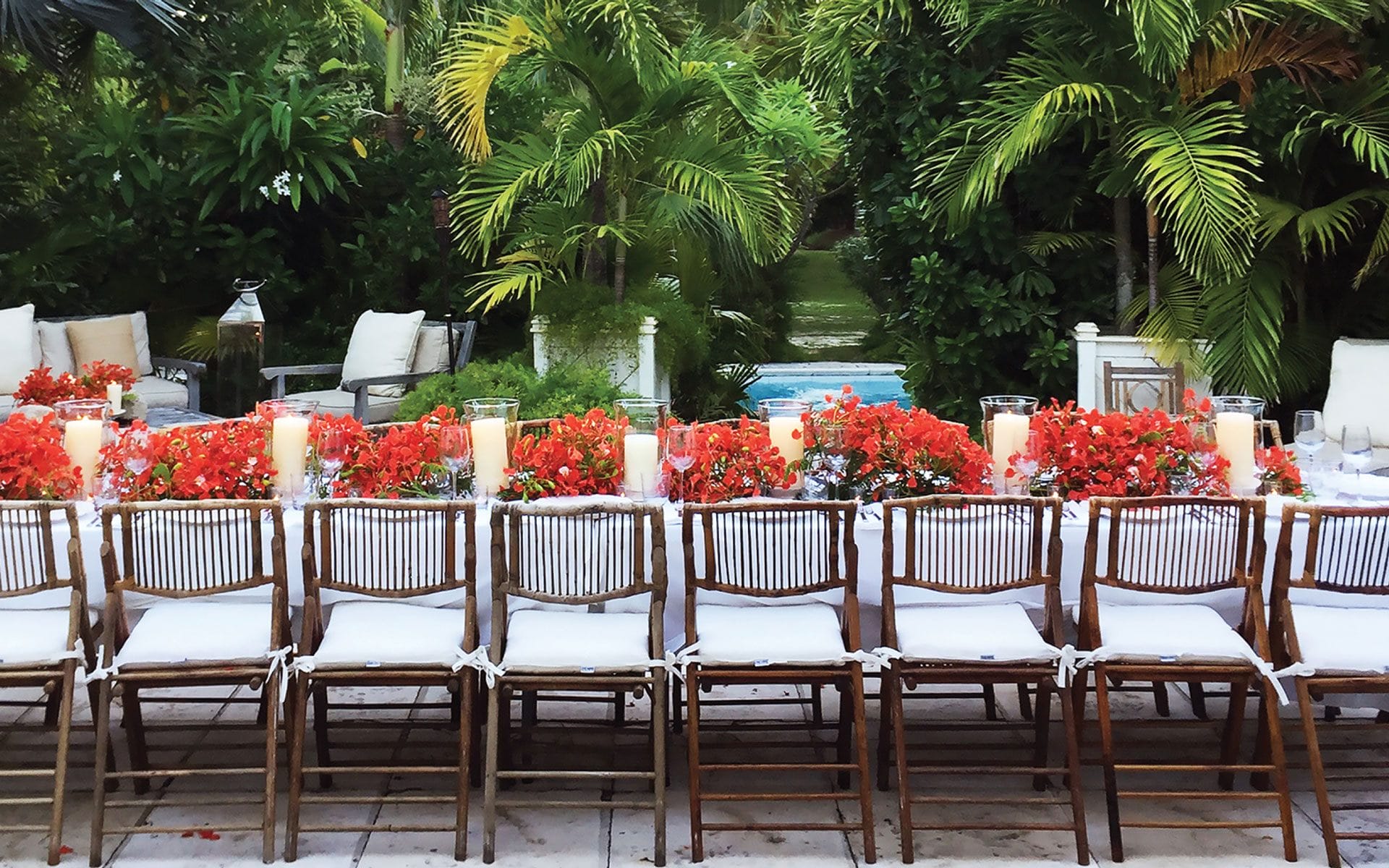 Outdoor table setup with red flowers, candles, and chairs surrounded by lush greenery.