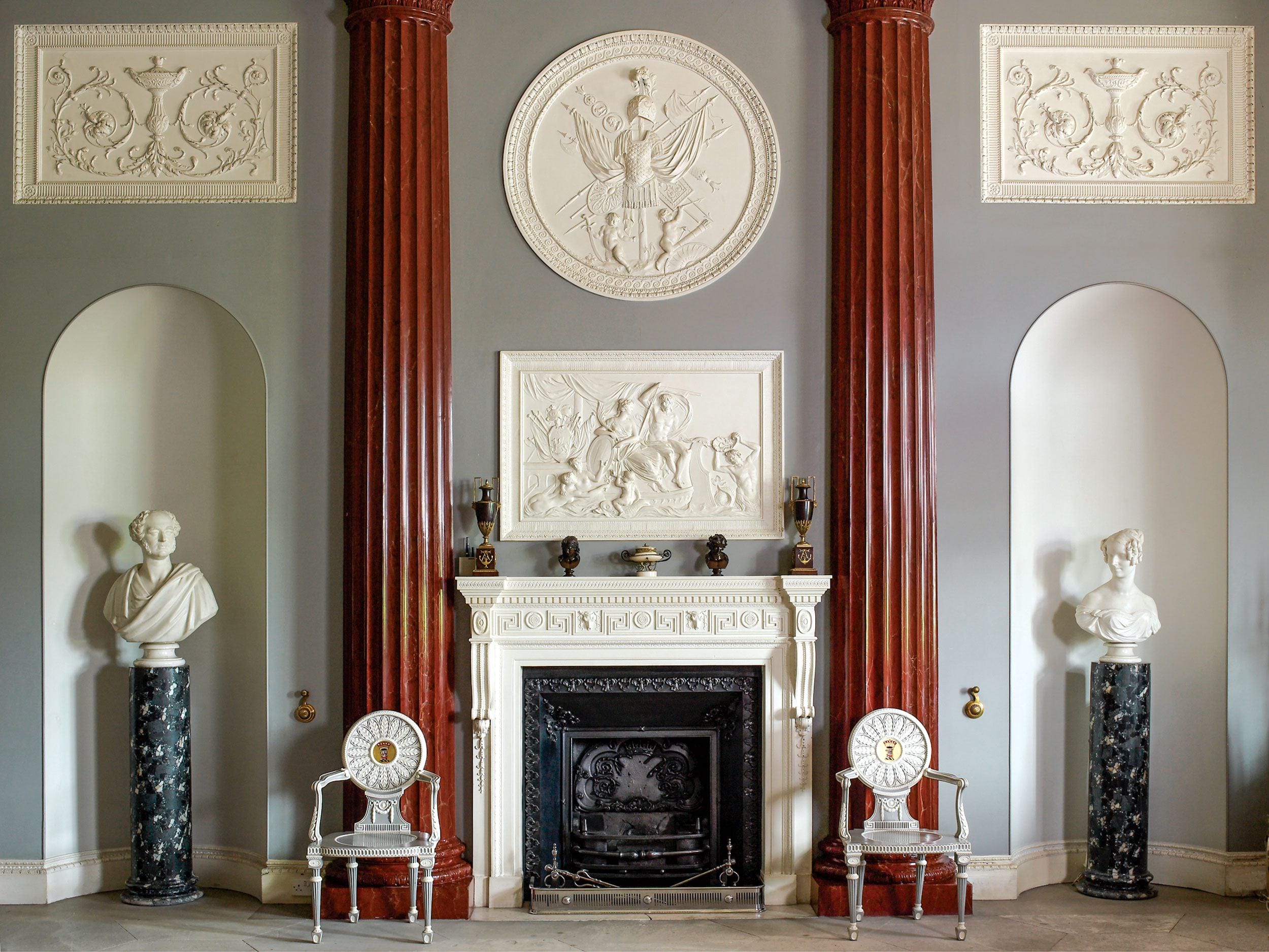 Elegant classical room with tall red columns, ornate fireplace, and busts on pedestals against a gray wall.