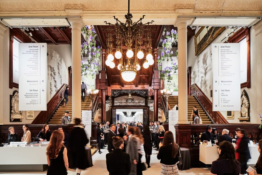 Visitors gather in a grand hall with chandeliers and ornate woodwork, engaging at a busy event with informational banners.