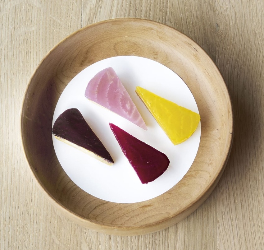 Four cheesecake slices with colorful toppings arranged on a white plate, placed in a round wooden bowl on a wooden surface.