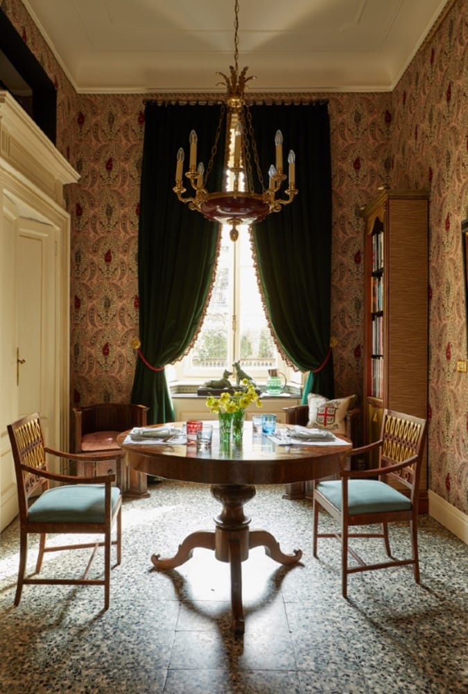 Elegant dining room with ornate chandelier, round wooden table, vintage chairs, and patterned wallpaper near large window.
