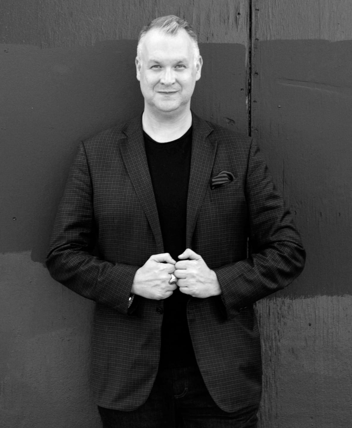 Man in a dark blazer stands smiling against a textured wall background in black and white.
