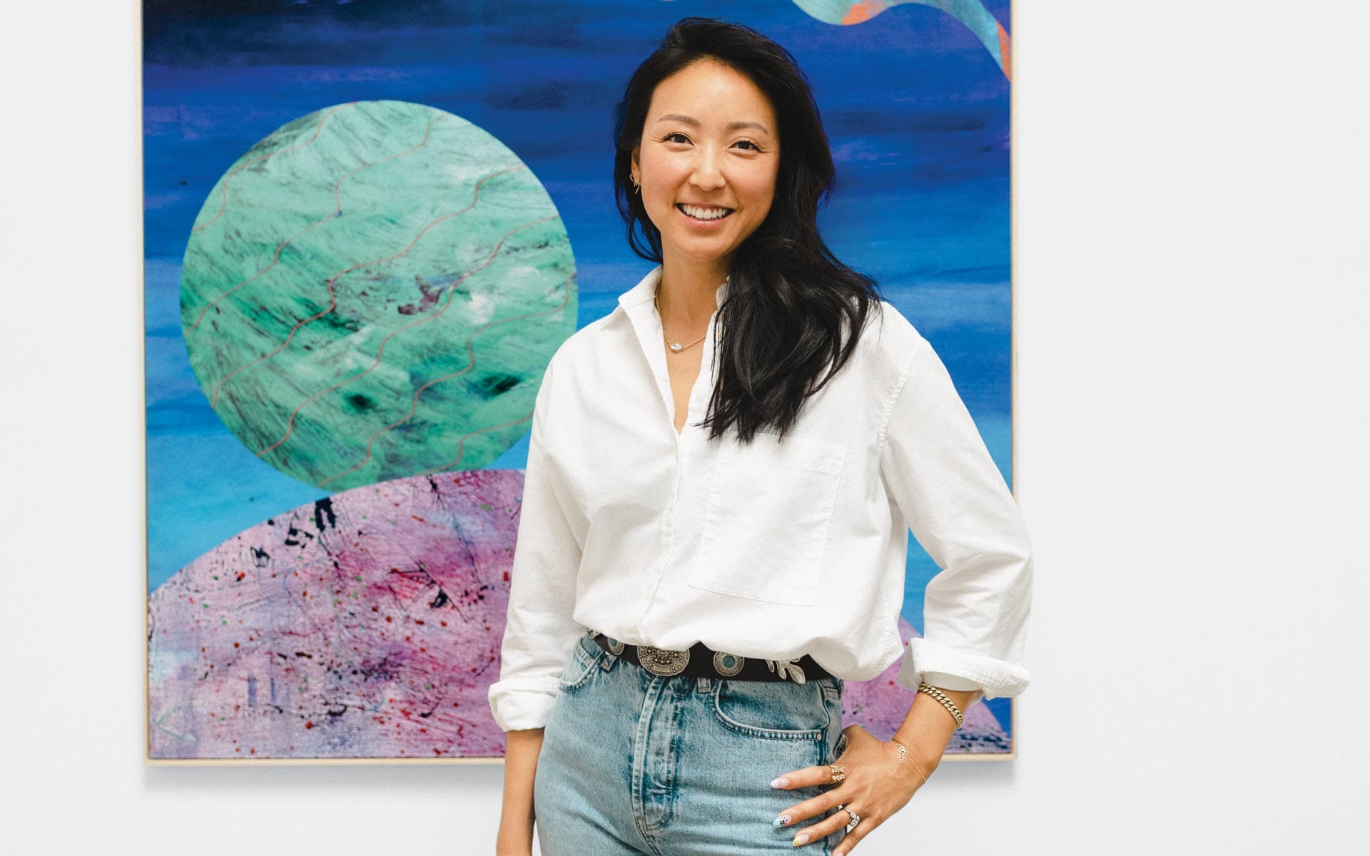 Person standing in front of colorful abstract artwork, wearing a white shirt and jeans, smiling at the camera.