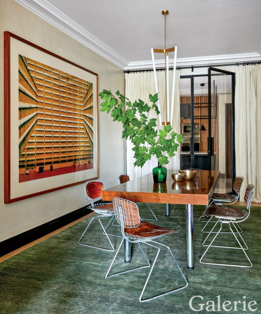 Modern dining room with wooden table, wire chairs, abstract art, lush plant centerpiece, and large window with curtains.