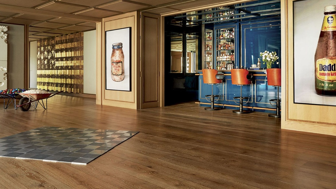 Modern art gallery with large bottle artwork, a polished wooden floor, and a view into a blue-themed bar with orange barstools.
