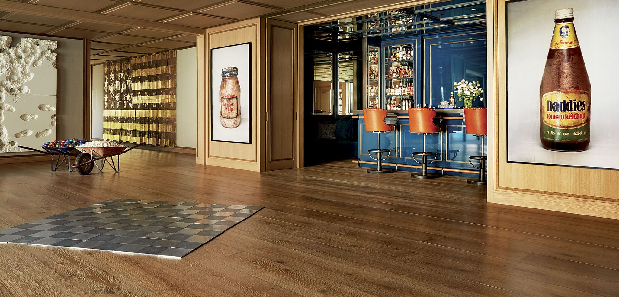 Modern art gallery with large bottle artwork, a polished wooden floor, and a view into a blue-themed bar with orange barstools.