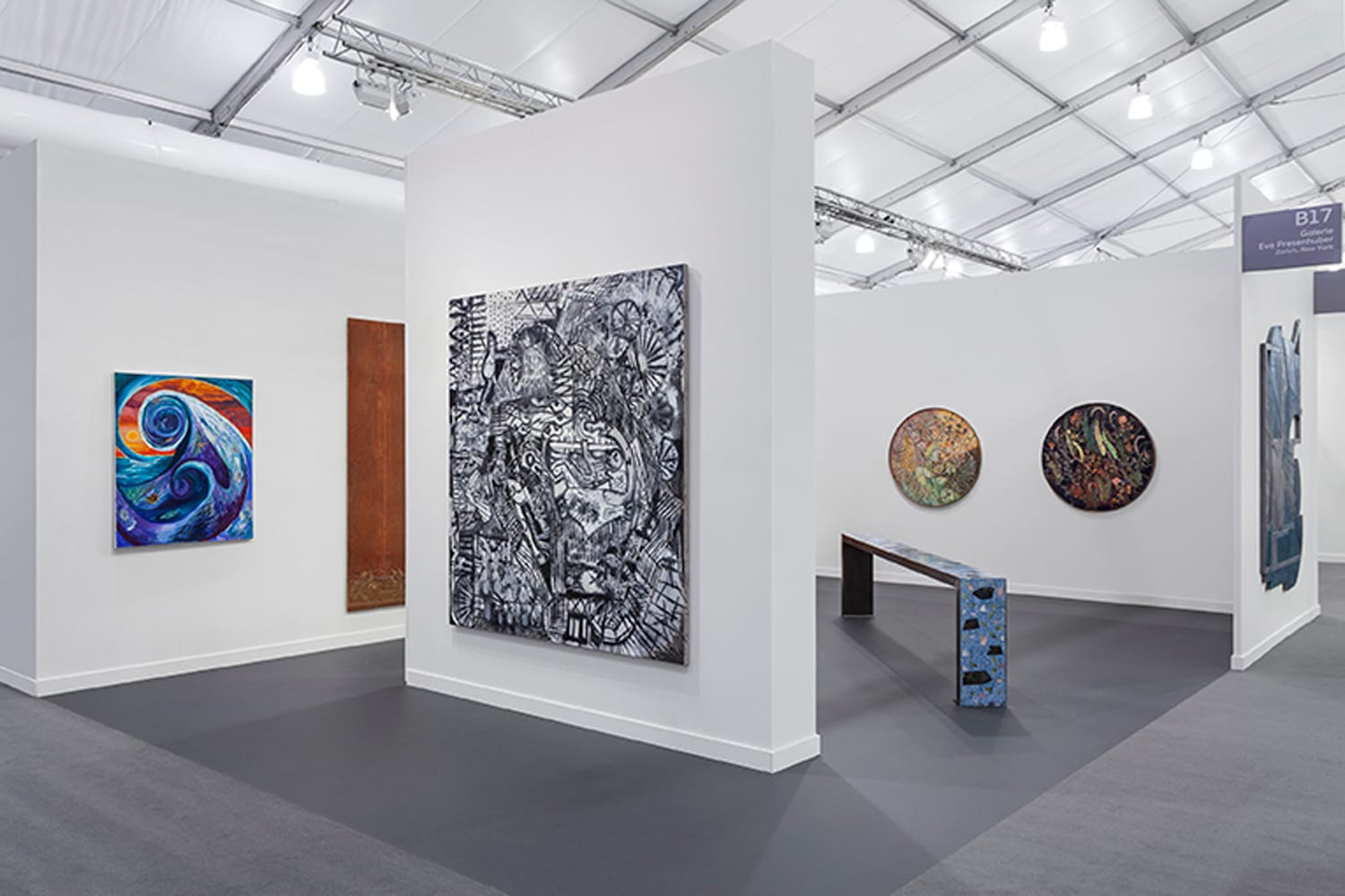 Art gallery exhibition with various colorful paintings and sculptures displayed on white walls and grey carpet flooring.