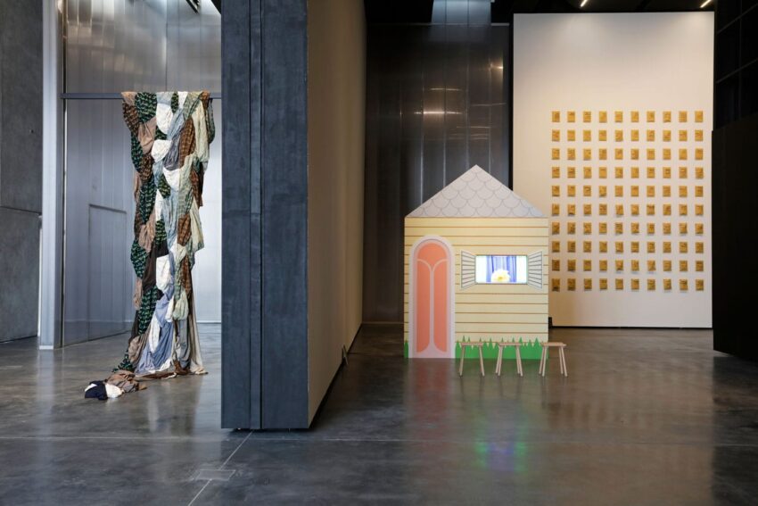 Modern art exhibit with a colorful house model, draped fabric installation, and grid of golden squares on a gallery wall.
