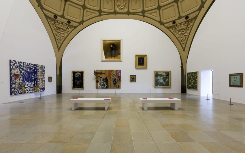 Art gallery interior with various framed paintings on white walls, arched ceiling, and two white benches in the center.