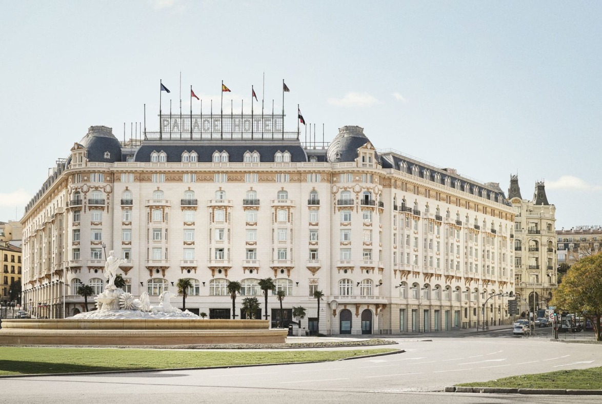 Large historic hotel building with ornate architecture, flags on the roof, situated beside a fountain and open plaza.
