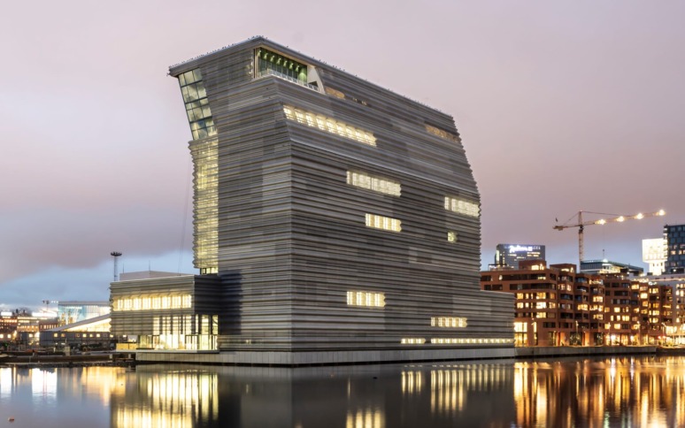Modern waterfront building with illuminated windows reflecting on calm water during twilight.