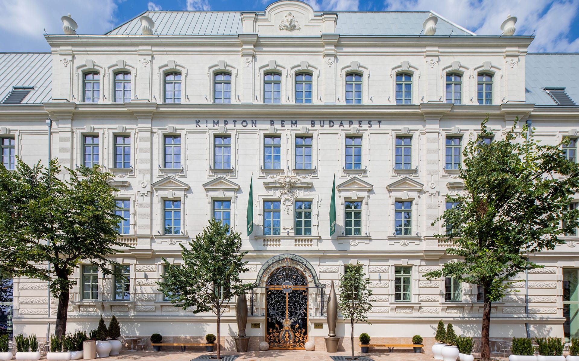 Front view of Kimpton Budapest, a classic white building with ornate architecture, trees, and a decorative entrance.