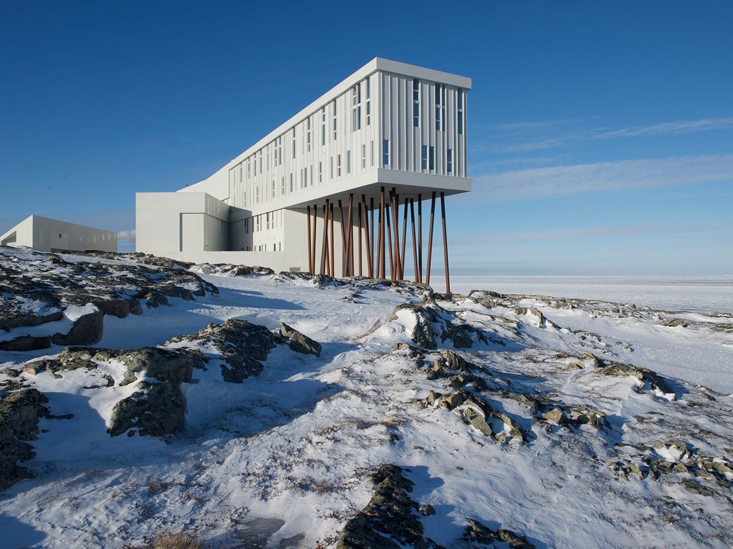 Modern building on stilts standing in snowy landscape with blue sky in the background.