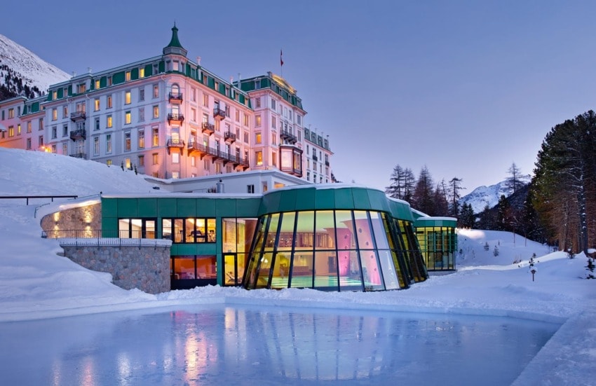 Historic hotel with modern glass extension surrounded by snow-covered landscape and mountains at dusk.