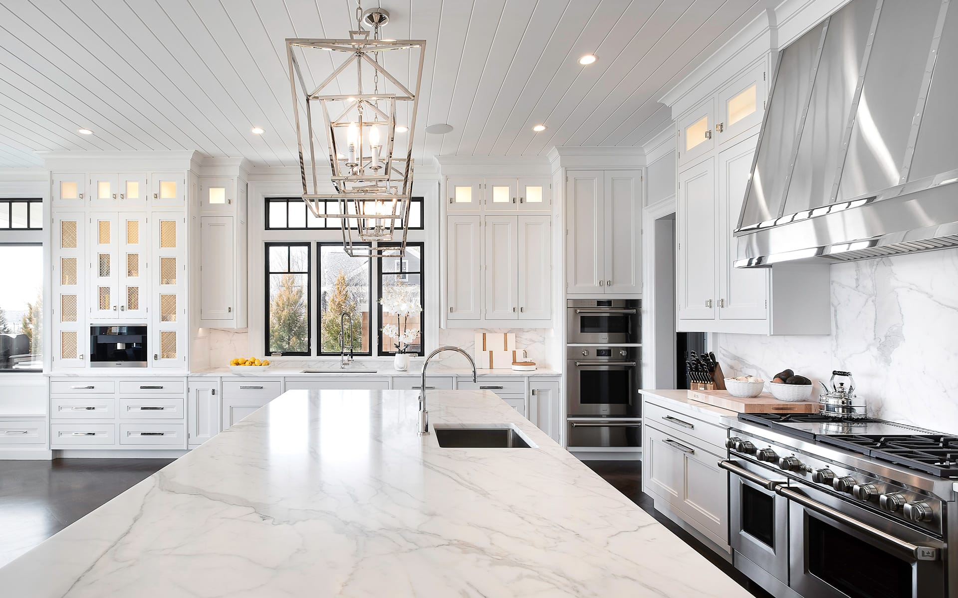 Modern white kitchen with marble island, stainless steel appliances, pendant lighting, and large windows for natural light.