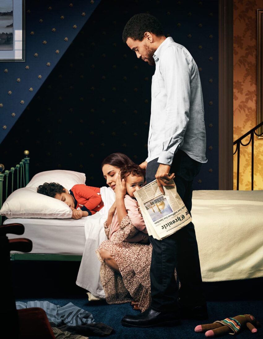 Man holding newspaper stands beside woman comforting children in a dimly lit bedroom environment.