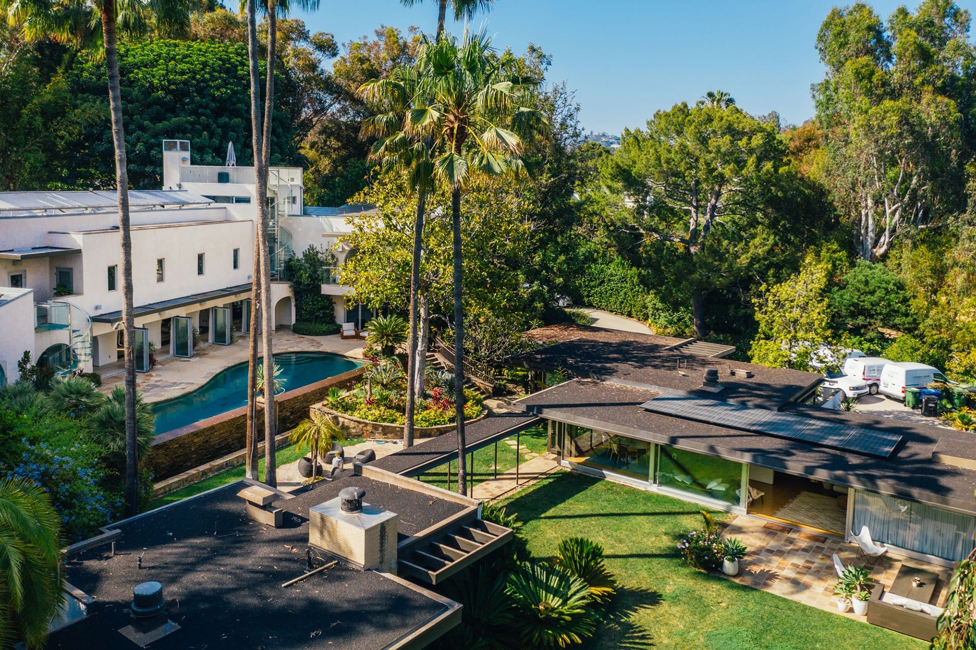 Aerial view of a luxurious estate with a swimming pool, surrounded by lush greenery and palm trees under a clear blue sky.