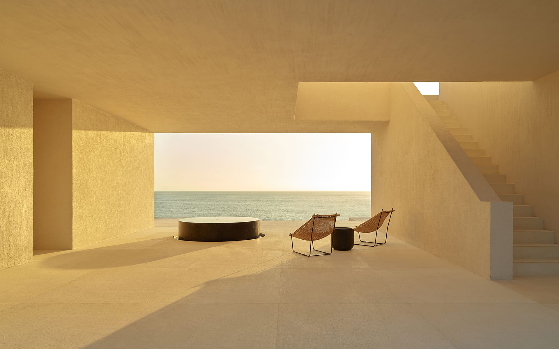 Minimalist terrace with two lounge chairs and a round table overlooking the ocean under a warm, golden sunlight.