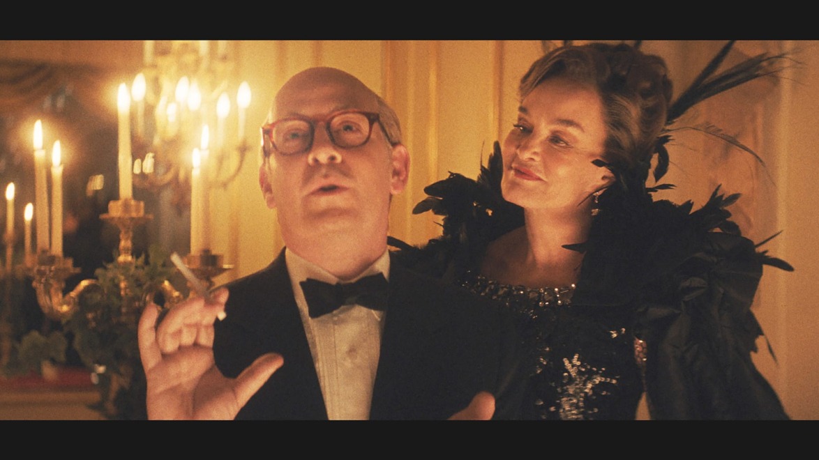 Man in tuxedo holding cigarette with woman in elegant dress, both at a candlelit event with warm ambiance