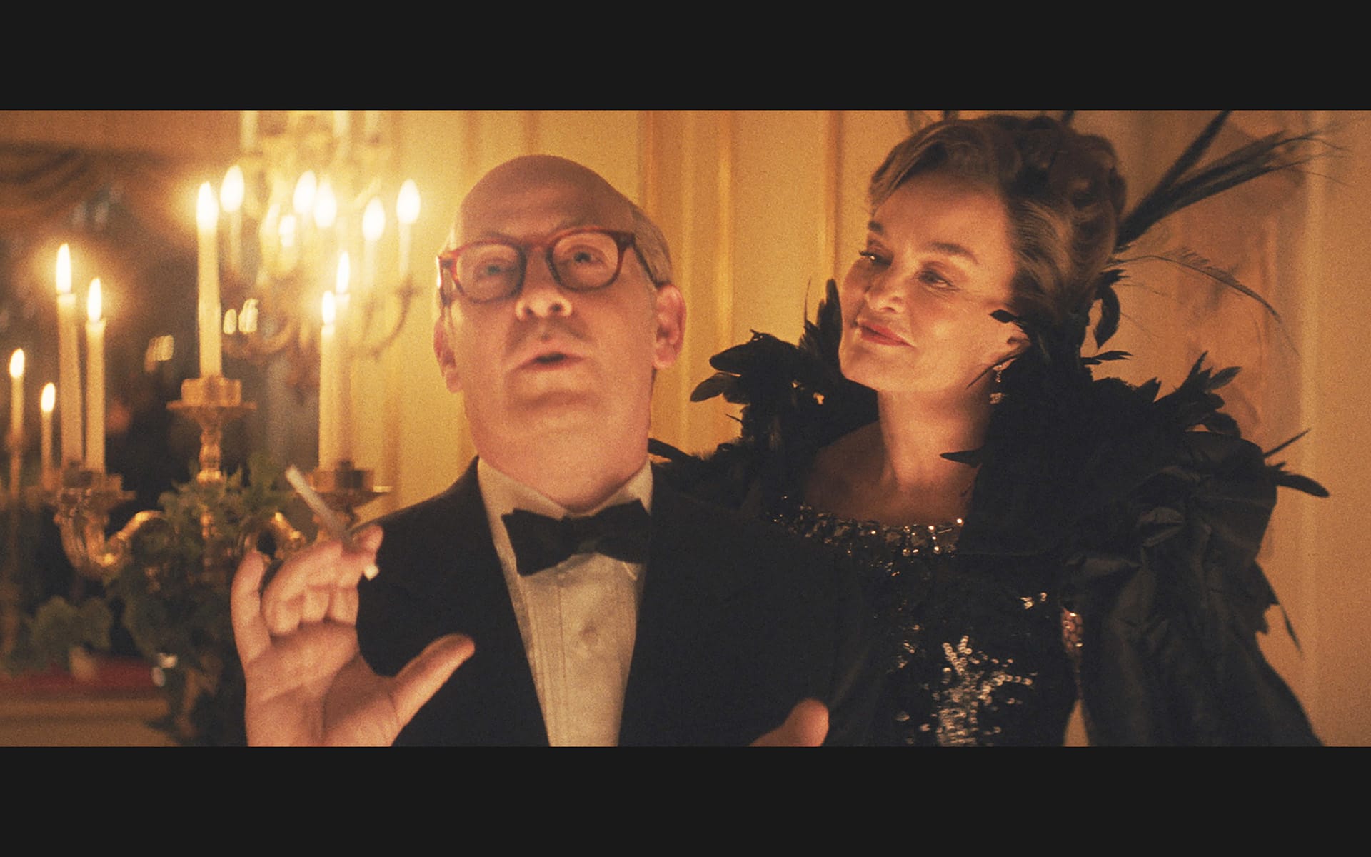 Man in tuxedo holding cigarette with woman in elegant dress, both at a candlelit event with warm ambiance