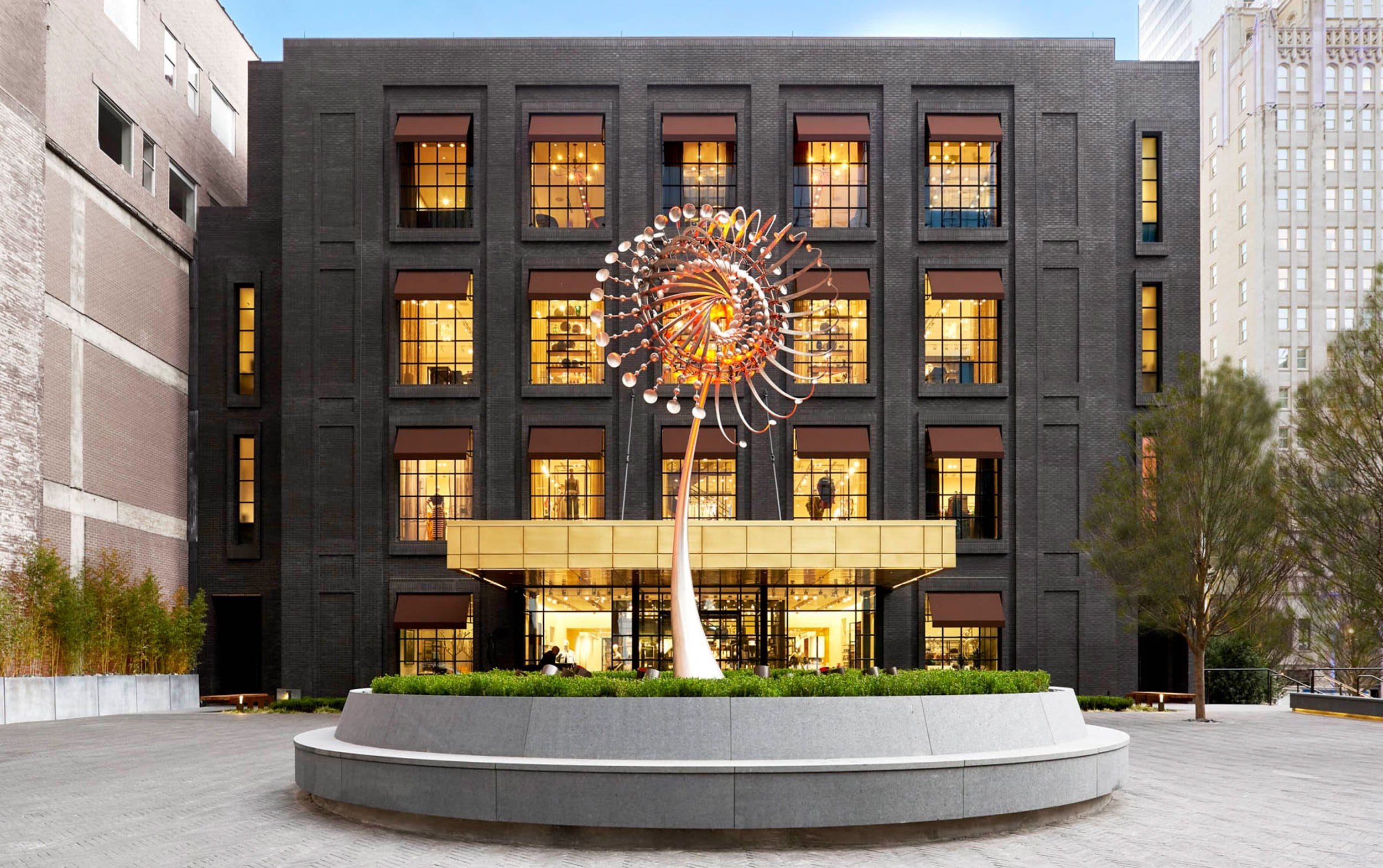 Modern building facade with large metal sculpture in front, surrounded by greenery and lit windows at dusk.