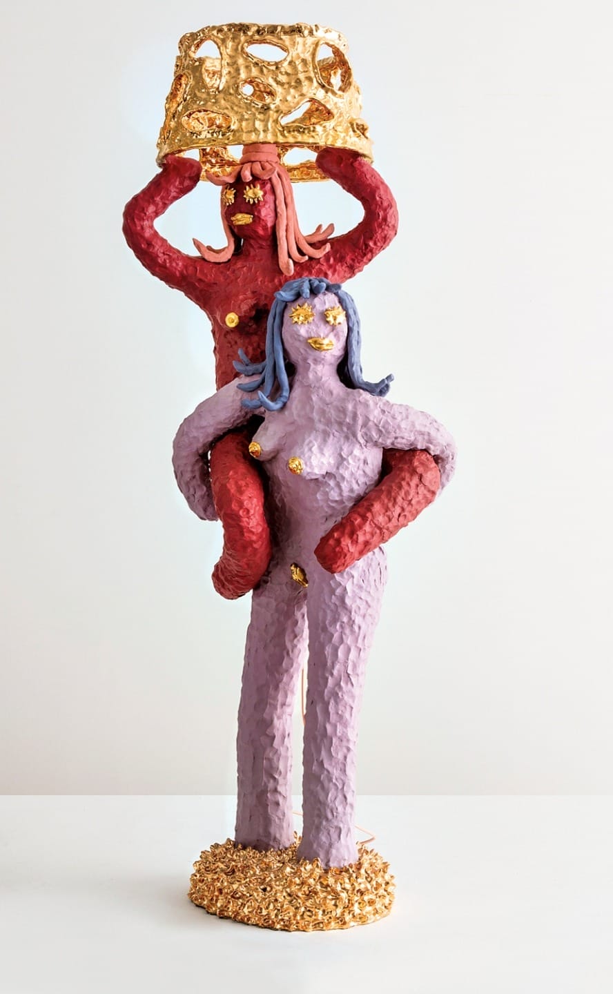 Two colorful, abstract human-like sculptures stacked, one with arms raised holding a gold object, set against a plain background.