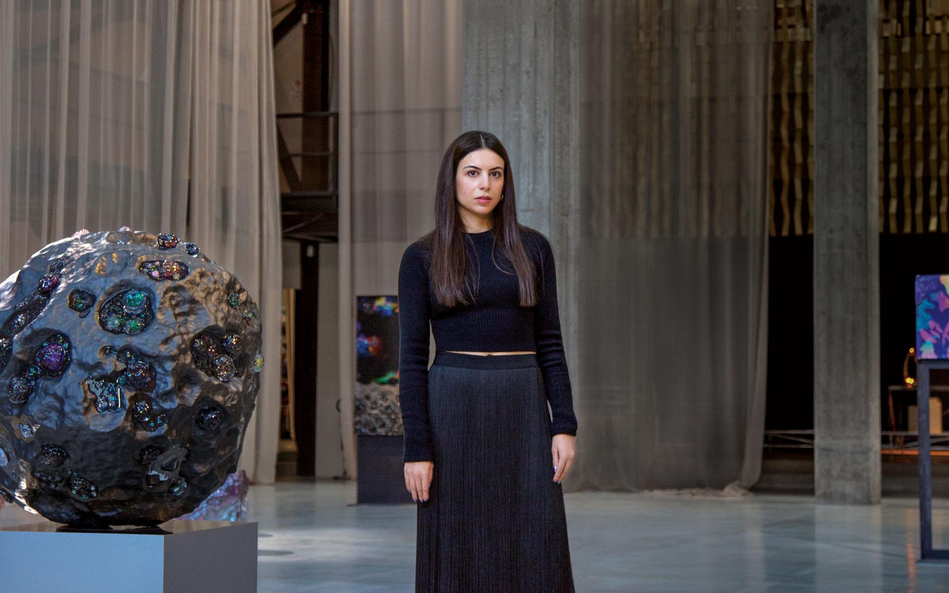 Woman in a black outfit stands in an art gallery with a large spherical sculpture on display.
